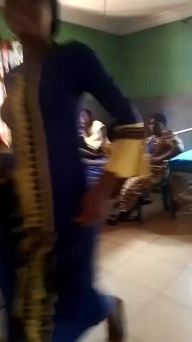 The video shows the interior of what appears to be a hospital room. The walls are painted green and white, and the floor is tiled. Two hospital beds are visible, with one patient lying on one bed connected to an IV drip. A woman sits on a blue plastic chair next to the bed with the patient. Another woman is sitting on another bed, interacting with a small child. A woman sits on a chair next to that bed. A person in blue clothing can be seen in the beginning of the video.