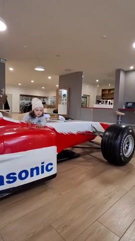 A little girl in a grey long-sleeved shirt and a striped beanie sits in the cockpit of a red and white Formula 1 race car replica. The car has "Panasonic" printed on its side. The girl's hands are on the steering wheel, which has many colorful buttons. She looks at the steering wheel. The scene takes place indoors, likely in a showroom or museum, with a polished wooden floor and displays in the background.