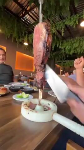 A man is carving a large piece of meat on a vertical metal skewer at a restaurant. The meat is dark brown and appears to be grilled or roasted. The skewer is held in a white plastic stand on a wooden table. He is using a large knife with a white handle to slice the meat. The man is wearing a striped shirt. There are other people seated at the table and in the background. The restaurant has a modern design with greenery hanging from the ceiling. There are plates and other items on the table, including limes. The lighting is bright, and the scene appears to be indoors.