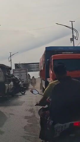 A view from a moving vehicle shows a busy road with several motorcycles and trucks. The rider of a motorcycle in the foreground is wearing a black shirt and a head covering. The road is dusty, and there is a large orange truck with a blue and red trailer to the right. In the background, there is a bridge under construction or repair, and other vehicles are visible. The sky is cloudy.