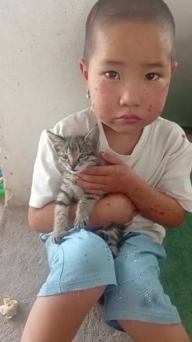 A young child, likely a girl given the hairstyle and clothing, sits outdoors holding a small, tabby kitten. The child has dark hair, fair skin with freckles and rosy cheeks, and dark eyes. She is wearing a white t-shirt and light blue shorts adorned with small white decorations. The kitten is held securely in the child's arms, with its front paws resting on the child's leg.

The setting appears to be a simple outdoor space, possibly a courtyard or a rural area, suggested by the concrete ground and a white, rough-textured wall in the background. To the left of the child, there's a glimpse of a green object and some debris on the ground. The lighting suggests it's daytime, and the overall atmosphere feels informal and candid.

The child is looking towards the camera with a gentle smile, and her expression conveys a sense of affection and contentment towards the kitten. The kitten, in contrast, looks a bit wary or curious, its ears perked and eyes alert.

A notable detail is a slight tilt of the child's head and a hand raised in a gesture that could be a peace sign or a wave, adding to the playful and engaging nature of the image. There is no visible text in the frame. The scene captures a tender moment between a child and a pet, showcasing innocence and simple joy. The location context of Chon-Kara, Kyrgyzstan, implies a rural or semi-rural setting consistent with the visual cues.