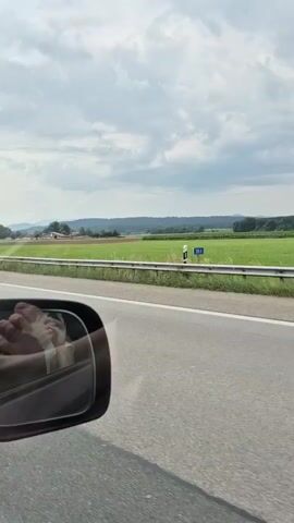 A side view mirror of a car shows a person's feet. The car is moving on a highway with green fields and some buildings in the background. A large blue truck drives past the car.