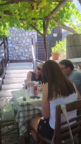 A group of people are gathered around tables in an outdoor restaurant setting. The tables are covered with checkered tablecloths, and there are glasses, bottles, and plates on them. Some people are sitting at the tables, while others are standing and talking. The restaurant appears to be located in a rustic setting, with stone walls and a grape vine-covered trellis overhead. Sunlight filters through the leaves, creating a dappled effect on the ground. There are also stairs visible in the background, leading up to another level of the restaurant. The overall atmosphere seems relaxed and casual.