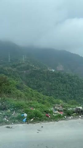 The video captures a drive through a mountainous region under an overcast sky. Lush green mountains are visible, with mist and low clouds partially obscuring their peaks. Several transmission towers are perched on the hillsides. The roadside in the foreground shows patches of green vegetation interspersed with litter and discarded items.

As the vehicle moves, a yellow road sign comes into view, displaying a speed limit of "30" in a red circle and text in Nepali and English. The English text partially reads "Under construction" and "Nagardunga - Na...". Further along the road, several makeshift shops or stalls constructed from corrugated metal sheets line the roadside, some featuring Coca-Cola and Pepsi branding. Goods are displayed for sale outside these stalls. A white truck is parked beside the road. The video concludes with a view of the winding road ahead, with an overhead metal structure spanning across. The scene depicts a journey through a scenic, hilly area, likely in Chandragiri, Nepal, given the location context and visible text.
