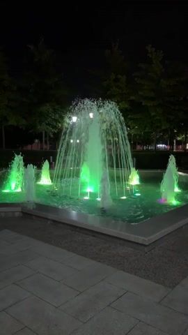 This is a vertical video of a fountain at night. The fountain has multiple streams of water spraying upwards and outwards. The water is illuminated by colored lights that change from blue to pink to green. The fountain is surrounded by a square basin with a stone border. In the background, there are trees with dark leaves and a dark sky. The ground in front of the fountain is paved with light-colored square tiles. The video shows the lights on the fountain changing color over a short period of time. There are no people or animals visible in the video.
