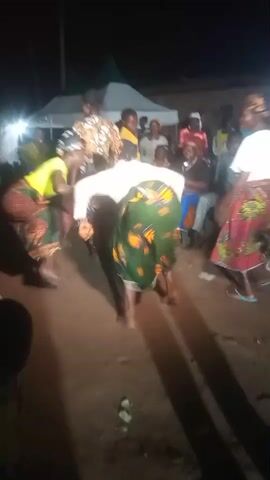 The video shows a night-time outdoor gathering. Several people are visible, with a focus on a group of women dancing in the center. They are wearing colorful clothing, including head wraps and patterned skirts. Some are bent over, possibly in rhythm with music or a specific dance move. In the background, there are tents and other individuals observing the activity. The ground appears to be dirt, with some debris scattered around. Lighting appears to be artificial, possibly from lamps or flashlights, creating shadows.
