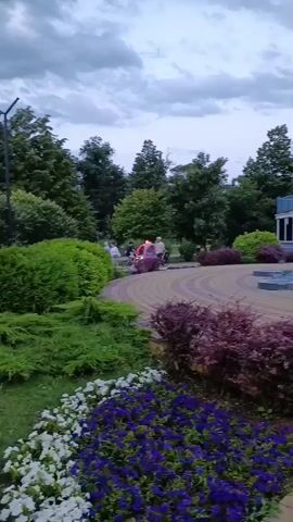 The video opens with a view of a park area. A fountain with pink and blue lights is visible in the center of the frame. To the left of the fountain, there is a structure with glass windows. Further to the left, a Ferris wheel can be seen. In the foreground, there are flower beds with purple and white flowers, and some shrubs. The ground is paved with patterned bricks. In the background, there are trees and a cloudy sky. Two people on bicycles are visible in the distance, near a vehicle with red lights. As the video progresses, the camera pans slightly to the right, showing more of the fountain and the surrounding park. The fountain's lights create a colorful display. The sky remains cloudy throughout the video. The overall atmosphere appears to be serene and peaceful, possibly in the late afternoon or early evening.