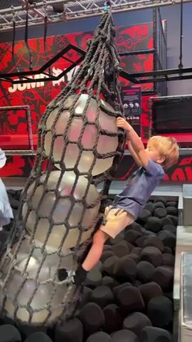 The video shows two small children trying to climb on a rope net containing three large balls. The net hangs over a pit filled with black foam blocks. The setting appears to be an indoor playground or activity center, with a background featuring red and black graphics and the word "JUMPYARD."

One child, a young boy wearing a blue shirt and khaki shorts, is seen briefly hanging onto the side of the net. Another child, a young girl wearing a white dress and leggings, attempts to climb into the net. The camera remains stationary, focusing on the children and the rope net.