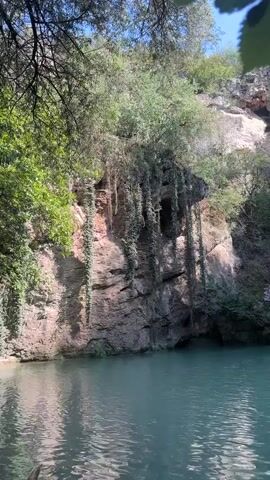The video shows a serene natural scene of a turquoise body of water in the foreground, likely a lake or calm river, with a rocky cliff face rising behind it. Lush green foliage, including trees and vines, cascades down the cliff, creating a beautiful, wild atmosphere. A waterfall can be seen cascading down the rock face on the right side of the frame, feeding into the water below. Sunlight filters through the trees, casting dappled light on the scene. The overall impression is one of natural beauty and tranquility.