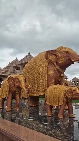The video opens with a close-up of three large, ornate, golden elephant statues, two smaller ones in the foreground and a larger one behind them. The elephants are covered in intricate patterns and have bells around their necks. The background reveals a detailed, multi-tiered white temple with ornate carvings and flags on top. As the video progresses, the camera pans to the right, showing a group of people gathered by a reflective pool of water in front of the temple. Several individuals are leaning over the edge of the pool, with some touching the water. The sky is overcast.