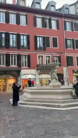 The video shows a square in an old European town. The square is paved with cobblestones and surrounded by buildings of various colors, all three to four stories high, with window shutters. There's a fountain in the foreground and people are walking around and sitting at outdoor cafes. A guitarist is preparing to play on the side of the square. There are various shops with lit interiors around the square. The sky is overcast.