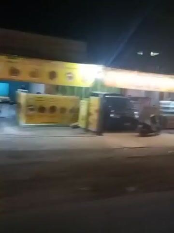 The video shows a nighttime view of a street with several businesses and parked vehicles. In the foreground, a dark-colored car and a motorcycle are visible. The businesses appear to be shops or restaurants, illuminated by bright signs and lights. One shop has a prominent yellow sign and a green awning. Another building is decorated with string lights. Further down the street, there are more shops, some with signs in a language that is not English. Several motorcycles are parked in front of these establishments. The overall scene suggests a lively commercial area at night. The video is somewhat shaky, indicating it was likely filmed from a moving vehicle.