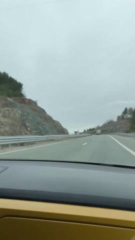 A car is driving down a highway on an overcast day. The road is surrounded by rocky cliffs on either side. There are a few trucks in the distance ahead. The car's dashboard is visible, and it is a light brown or tan color. The sky is grey and cloudy.