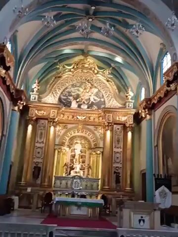 The video shows the interior of a church. The camera pans across the space, focusing on the altar area, which is ornate with gold details and religious iconography. There are paintings on the walls, and a round window lets in natural light. The ceiling is vaulted and decorated with chandeliers. There is no discernible human activity. The overall impression is one of grandeur and religious reverence.