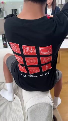 The video shows a close-up of the back of a person wearing a black long-sleeved shirt. The shirt has a graphic design consisting of nine red squares arranged in a 3x3 grid, with white line drawings inside each square. The drawings depict a camera, a bicycle, a computer, cans of soda, an "@" symbol, headphones, a video game controller, a glass of drink with coffee beans, and a lighter. Below the grid, the text "The station of ad." is printed in white. The person is sitting on a sofa, with their legs crossed. The background is blurred but appears to be an indoor setting with kitchen elements visible. Another person is partially visible in the background, holding a glass. The video is short, approximately 5 seconds long, and is zoomed in on the shirt.