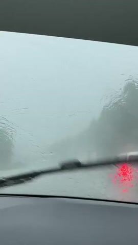 The video shows a view from inside a vehicle through a rain-streaked windshield. Water droplets and streaks distort the vision of the outside scene. In the distance, through the haze and rain, a red light is visible, likely from another vehicle's taillight. The overall scene is washed out and gray, indicating overcast or stormy weather, possibly during the daytime due to the limited visibility. The interior of the car is visible at the bottom of the frame, appearing to be a dark dashboard. The location context provided is Kliuchevsk, Russia. The activity is driving in heavy rain or fog, which severely limits visibility. The emotional tone conveyed is one of reduced visibility and potentially cautious driving due to the weather conditions. There is no visible text in the media file.