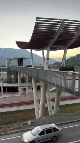The video pans across a modern architectural structure with a curved roof supported by pillars. A elevated walkway with metal railings extends from the structure. Below the walkway, a person rides a bicycle on a red track. In the background, mountains covered in trees are visible, with a clear sky above. A road runs in the foreground, and a car is partially visible on the left side. The overall scene appears to be set during either sunrise or sunset, indicated by the soft lighting and the gradient of colors in the sky.