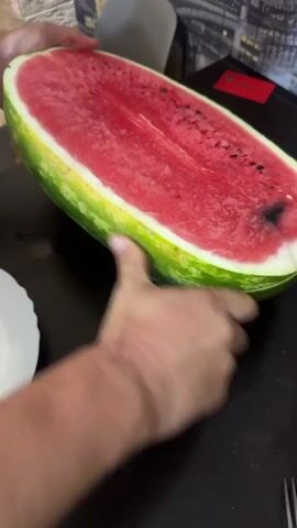 In this media file, a person is cutting a large, ripe watermelon in half. The watermelon is oval-shaped with a green rind and bright red flesh, speckled with black seeds. The cut surface reveals the juicy interior. The person's hands are visible, one holding the watermelon steady while the other wields a knife. A white plate is placed to the left of the watermelon, presumably to catch the cut pieces.

The scene appears to be indoors, likely in a home or kitchen, given the dark table and the background which shows a portion of a patterned chair and a hint of what might be a cityscape or building facade in the distance. The lighting suggests it could be daytime, but the specific time of day or weather conditions are not discernible.

The activity is straightforward – preparing to serve or eat the watermelon. There are no strong emotional cues conveyed, as the focus is on the action of cutting. No visible text is present in the frame. The context provided places this event in Sverdlovskiy, Russia.
