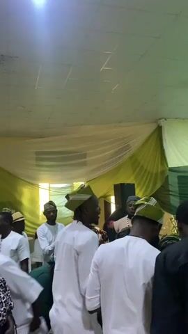 The scene captures a vibrant social gathering, likely a celebration or event, indoors. Several people are visible, with the men primarily dressed in white tops, some paired with traditional green hats. Other attendees wear varied outfits, including a man in a traditional patterned top and a woman in a green headwrap. The room is decorated with white and green drapes hanging from the ceiling. Music plays in the background, and the atmosphere is lively, with people interacting and moving about. A window is visible, letting in natural light.