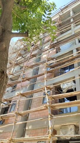 The video shows the exterior of a multi-story building under construction. The building is primarily brick, with concrete sections and window frames. The facade is covered in wooden scaffolding that obscures the building's details. The scaffolding is constructed using thin poles and planks tied together. The sky is visible in the background, indicating a sunny day. A tree with green leaves partially obscures the lower left corner of the frame. There are no people visible in the video.