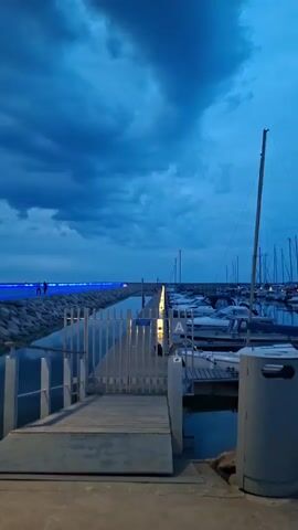 The video shows a serene waterfront scene at dusk in Tallinn, Estonia. The sky is filled with dramatic, dark blue clouds, suggesting an approaching storm or the remnants of one. The setting is a modern marina with several boats docked and illuminated by soft yellow lights. A concrete walkway extends into the water, lined with rocky barriers and bathed in a vibrant blue light, creating a striking contrast with the muted colors of the sky and sea.

Several individuals are visible on the walkway and near the marina, appearing to be enjoying the evening ambiance. Some are walking, while others stand in small groups, possibly engaged in conversation. The presence of streetlights, one prominently featured in the foreground, indicates that the scene is transitioning from daylight to night. The overall mood is peaceful and contemplative, enhanced by the twilight glow and the rhythmic lapping of water against the boats. The natural beauty of the cloudy sky and the man-made elements of the marina blend harmoniously to create a captivating view.