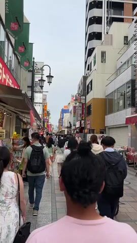 A vertical shot captures a busy pedestrian street in Japan. The perspective is from behind a person wearing a pink t-shirt, walking with a group of people. The street is lined with shops and tall buildings, many featuring signs with Japanese characters. The sky is overcast. People are walking in both directions, some carrying bags and backpacks. A bicycle is visible in the middle distance. The atmosphere appears to be that of a lively commercial area.