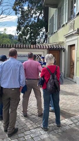 In an outdoor, paved area with cobblestones, a group of people observes a person in historical costume. Three adults are in the foreground with their backs mostly to the camera. The person on the left wears a blue and white plaid shirt and tan pants. The person in the middle wears a red and white checkered shirt and tan pants, holding what appears to be a small device or camera. The person on the right wears a red shirt and blue jeans with a patterned backpack. Another person, partially visible, stands behind the person in red.

From behind a white sign, a man dressed in an elaborate blue and white historical costume with ruffles and a feathered hat emerges, holding a halberd or staff. He stands facing the onlookers.

The setting includes a light green building with brown wooden doors and window shutters on the right. On the left, a white sign displays the text "buchwalder linder" and a partial phrase "möbel teppiche". Trees and a distant building on a hill are visible in the background under a partly cloudy sky. Shadows indicate bright daylight.