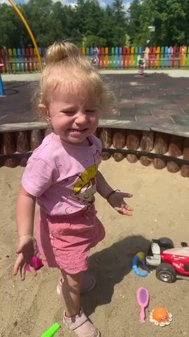 A young girl with blonde hair pulled up in a bun, wearing a pink t-shirt and pink shorts, stands in a sandbox. She is looking down at her hands, which are covered in sand. In front of her on the sand are several toys, including a red toy car, a purple shovel, and an orange toy with tentacles. Behind the girl is a wooden fence, and beyond that, a colorful playground fence. The ground appears to be covered in sand and wood chips. The lighting suggests it is daytime.