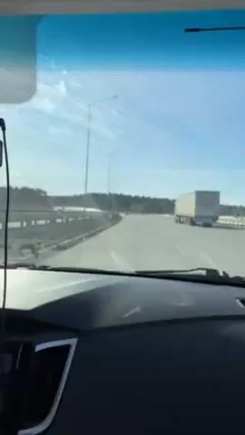 The video shows a view from inside a car driving on a highway on a sunny day. The windshield is a bit dirty. To the right, a large truck is driving ahead of the car. The highway is mostly empty, with a few other vehicles visible in the distance. On both sides of the road, there are trees and some patches of snow. There are several street lights lining the highway. The dashboard of the car is visible at the bottom of the frame. Music is playing in the background.