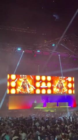 The video captures a live concert or event with a large audience. In the foreground, a densely packed crowd is visible, facing a stage. The stage features a massive LED screen displaying a vibrant graphic. This graphic alternates between a stylized image of a person, possibly a performer, wearing a golden headdress and surrounded by circular, glowing elements, and abstract patterns with warm colors like orange and yellow. Below the screen, a DJ or performer is subtly visible at a console, illuminated by green and blue lights. Above the stage, a complex lighting rig with numerous spotlights and trusses is suspended, creating beams of light that cut through the atmospheric haze. The overall ambiance is energetic and immersive, typical of a large-scale entertainment event.