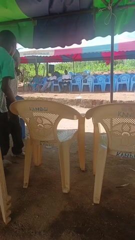 A person in a green shirt walks into the shot behind two tan plastic chairs that say “KANDU” on them. A child in a patterned shirt stands beside the chairs. Both individuals are under a tent with a blue and green canopy. Another tent with a red and white canopy sits in the background, and there are rows of empty blue plastic chairs behind that. The ground is brown dirt. A phone number is visible on the edge of the tent canopy in the background.