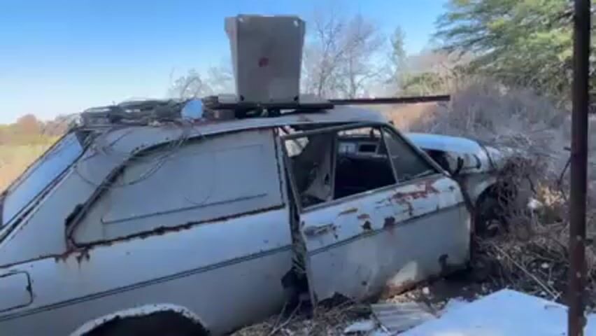 The video shows a dilapidated, light-colored hatchback car, likely abandoned in a rural, overgrown area. The car is rusted and in poor condition with an open door. There's a metal container or box strapped to the roof along with a jumble of wires. The surrounding environment includes dry grass, trees, and a clear blue sky. The landscape suggests a field or open area. The camera pans up from the car to showcase the surrounding scenery.