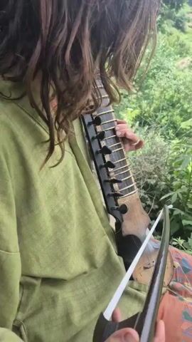 A person with long, wavy brown hair, wearing a green tunic and patterned pants, sits outdoors playing a stringed instrument. The instrument, held upright on their lap, has a dark fretboard with several strings stretched across it. The person's left hand is positioned on the fretboard, while their right hand holds a bow, drawing it across the strings to produce music. The background is lush and green, with foliage suggesting a natural, possibly hilly, environment. The lighting indicates it is daytime. The scene appears peaceful and focused on the act of playing music in a serene setting. There is no visible text in the media file.