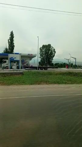 The video shows a highway scene with a gas station in the background. A white truck with a blue awning is parked at the gas pumps. A large red tanker truck is also visible. The sky is overcast and the weather appears to be cloudy. The road is paved and there is vegetation along the side. The camera is moving, suggesting it is mounted on a vehicle.