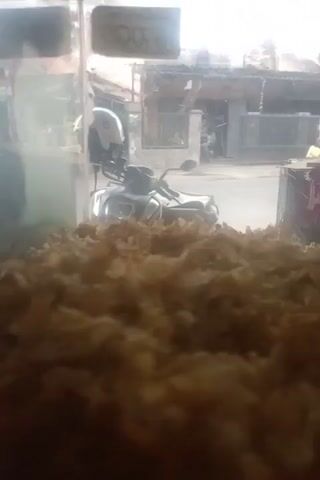 The video shows a close-up of what appears to be a terrarium or enclosure filled with wood shavings or a similar substrate. In the lower left corner, a dark, rounded object is partially visible, possibly an animal or a decorative item. Through the glass and the substrate, a blurry outdoor scene is visible, including what looks like a parked motorcycle and buildings in the background. The lighting is dim, and the focus is primarily on the substrate within the enclosure.