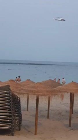 The video shows a beach scene with stacked lounge chairs on the left and thatched umbrellas in the foreground. In the middle ground, several people are visible in the calm blue water. The background consists of the sea meeting a hazy sky. A white and orange helicopter is flying in the upper right portion of the frame. The sky is overcast and gray.