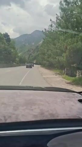 The video shows a car driving on a road with mountains in the background. There are trees on both sides of the road. Another car is ahead in the same direction. The sky is cloudy. Music plays. The dashboard of the camera's vehicle is visible in the foreground.