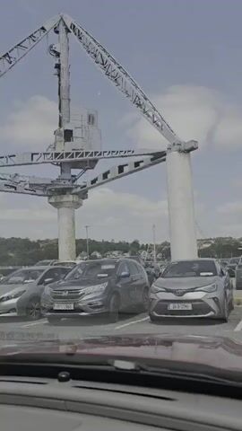 This media file captures a scene of a parking lot under a large, industrial structure, likely a crane or part of a port facility. Several cars are parked in the lot, including SUVs and sedans. The time of day appears to be daytime, with the sky partly cloudy. The weather seems fair.

In the background, there are trees and buildings, suggesting an urban or semi-urban environment. A few people are visible, standing and walking in the distance near the parked cars. The dominant feature of the scene is the imposing metal structure overhead, which is a complex assembly of beams and supports, with a cab-like structure at its apex. This structure creates a dramatic overhead visual element, contrasting with the more mundane setting of the parking lot.

License plates are visible on some of the cars, indicating they are registered in Ireland (e.g., "211-D-274", "231-D-3366"). The overall mood of the scene is neutral, capturing a moment in a functional, everyday location. The focus is on the juxtaposition of the industrial infrastructure with the everyday presence of vehicles and people. The quality of the media suggests it's a still frame taken from a video.