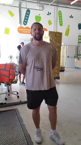 A man stands in what appears to be a warehouse or distribution center. He is wearing a light brown t-shirt with a graphic on it and dark shorts, along with white sneakers and socks. His hands are clasped behind his back. He is facing the camera and appears to be smiling slightly. In the background, there are stacks of yellow boxes on pallets. The walls are white and decorated with colorful graphics of food items such as bread, apples, and bottles. To the left of the man, there is a white chair with an orange safety vest draped over it, and behind that, another person is partially visible. The lighting suggests an indoor environment.