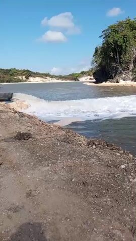 The video shows a natural landscape under a bright blue sky with white clouds. The foreground features a dry, sandy embankment with some scattered rocks and dirt. A body of water, appearing to be a river or inlet, dominates the midground and background. White foamy water is visible, suggesting some kind of current or outflow. On the right side of the frame, a cliff face with white rock and green vegetation rises above the water. The left side of the frame shows a more distant, lower shoreline with sand and vegetation. The scene is a natural outdoor environment with no people or man-made structures other than a visible pipe discharging into the water.