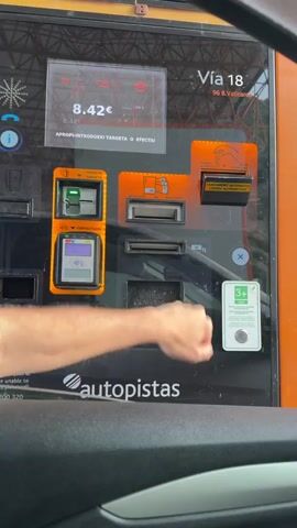 The video shows a close-up shot of a payment machine for a toll road. A person's arm and hand are visible in the foreground, interacting with the machine. The machine has various slots for inserting cash, cards, and tickets, as well as a display screen showing the amount due, which is 8.42€. The person's hand is seen touching a button on the right side of the machine, possibly to confirm a payment or to select an option. The environment appears to be outdoors, possibly at a toll booth, with a hint of greenery in the background. The machine itself is orange and black, with Spanish text indicating its functions.