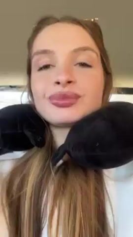 The video shows a light-skinned woman with long blonde hair wearing a white sweatshirt and a black neck pillow, talking directly to the camera inside a car. Her nails are painted red. She gestures with her hands as she speaks, and music plays in the background. The car interior is visible, including parts of the seats.