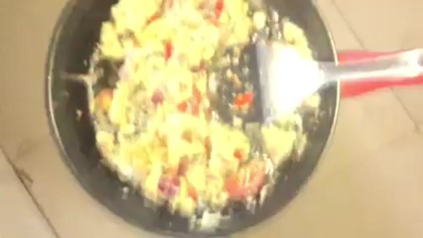 A top-down shot shows a black skillet containing scrambled eggs mixed with red onions, tomatoes, and other unidentifiable ingredients. The food is being stirred with a spatula, which has a black plastic head and a red handle. The skillet rests on a beige tiled surface. The image is blurry.
