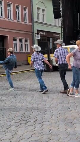 This is a medium shot, outdoors, of a line of people line dancing. They are on a cobblestone street in front of buildings. A yellow and white truck with a stage setup behind it is visible. The people are wearing cowboy hats and plaid shirts, and are dressed casually. The focus is on the line dancing action. The time stamp is 00:00.