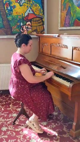 A woman in a red floral dress sits on a stool and plays a piano. She has her hair pulled back. She is wearing tan sandals. The piano is wooden and ornate. Behind the piano are two colorful paintings. The floor is covered with a red patterned carpet. Sunlight streams in from the left side of the frame.
