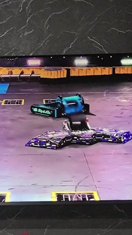 Two robotic combat vehicles are in an arena. One is blue and cylindrical with black accents and the words "Freak 37" visible on its side. The other is purple and flat with black accents and a complex design on its surface. The purple robot is positioned over the blue robot, and sparks are flying from the point of contact. A "REPLAY" button is visible in the lower left corner of the screen. The arena floor is purple with blue and yellow markings. The background consists of elevated yellow structures with lighting.