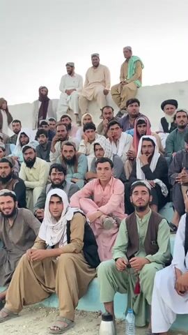 This video shows a group of men sitting in bleachers. Most of the men are wearing traditional clothing. The men are of various ages, and most appear to be looking towards the camera. Some of the men are holding prayer beads. The bleachers appear to be made of concrete. The background is a light-colored wall, possibly part of a stadium or outdoor arena. The sky is visible in the upper part of the frame and appears to be clear. The overall impression is that the men are attending an event or gathering.