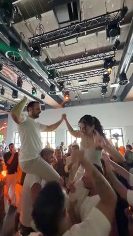 At a lively celebration, likely a wedding reception, a man is seated on the shoulders of another man, with a woman sitting on the shoulders of the first man. They are both smiling and holding hands, with their arms raised in the air. They appear to be dancing or being lifted by the crowd. Numerous people surround them, also with their arms raised, some clapping and cheering. The environment is an indoor venue with a high ceiling, featuring exposed beams, and numerous stage lights hanging from trusses. The lighting is a mix of bright overhead lights and colored stage lights, with a warm orange glow in some parts of the frame, suggesting a festive atmosphere.