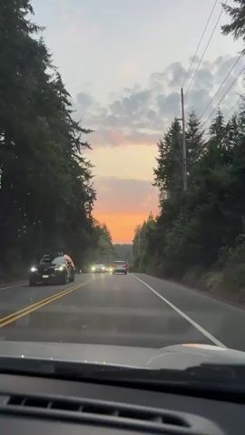 The video shows a point-of-view shot from inside a vehicle driving on a two-lane road lined with tall evergreen trees. The sun is setting, casting an orange and pink glow across the sky, with some clouds visible. Several cars are driving in the same direction as the viewer, with their headlights on. The road has a double yellow line in the center and white lines on the edges. There are utility poles visible on the right side of the road. The overall atmosphere is serene and peaceful, depicting a scenic drive during sunset.