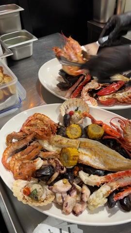 In a commercial kitchen setting, a chef with black gloves is preparing two large platters of seafood. The first platter is filled with various grilled seafood items including shrimp, fish fillets, corn on the cob, octopus, squid, mussels, scallops, and crab legs. The second platter contains a similar assortment, including grilled shrimp, lobster, crab legs, fish, and mussels. The chef uses a metal spatula to transfer additional grilled shrimp from a metal baking tray onto both platters. The kitchen environment includes stainless steel countertops, containers, and other cooking equipment.